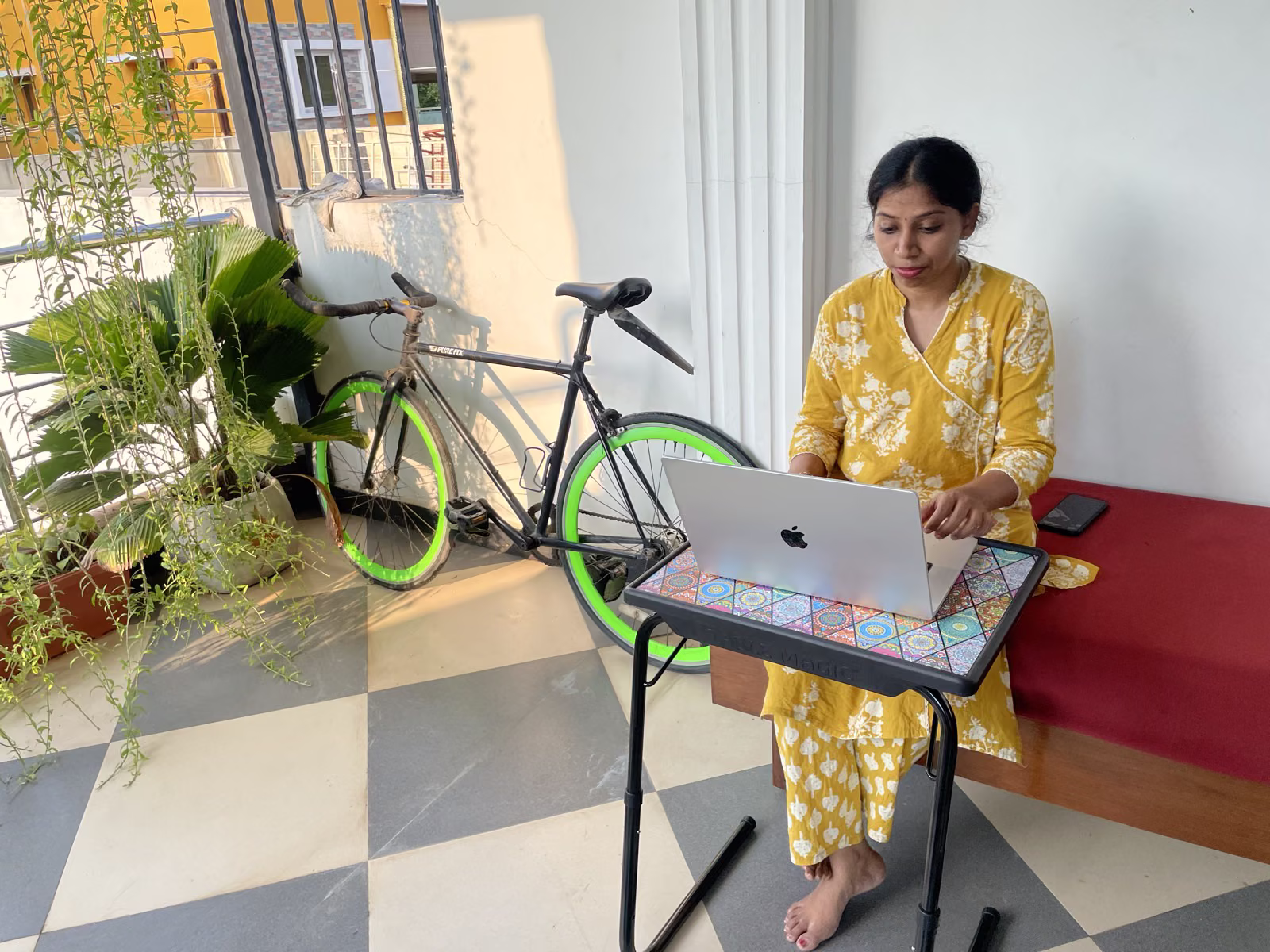 Guest working on her MacBook on the balcony at Nivaa Stays — adjustable stand, bicycle in the background